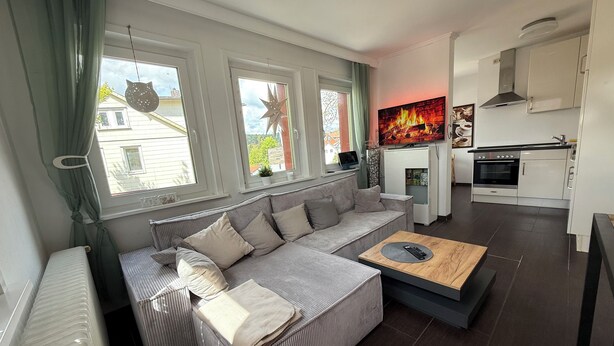 A modern living room with a grey sofa, a coffee table, and a TV. There is a kitchen area with a stove and cabinets. - PrimeLoft Eule - up to 10 pers - 2 floors - balcony - modern & stylish - central (Braunlage)