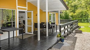 A covered outdoor area with a wooden deck, a seating area with tables and chairs, and a view of a green landscape.