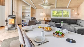Un salon chaleureux avec un plafond en bois, un poêle à bois encastré, une table à manger dressée avec des assiettes, des tasses et un panier de fruits.