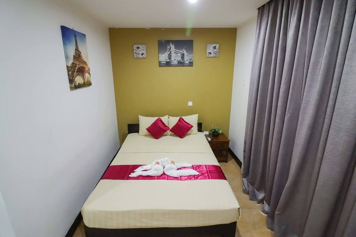 A hotel room with a bed, two pillows, a nightstand, a lamp, and a curtain.