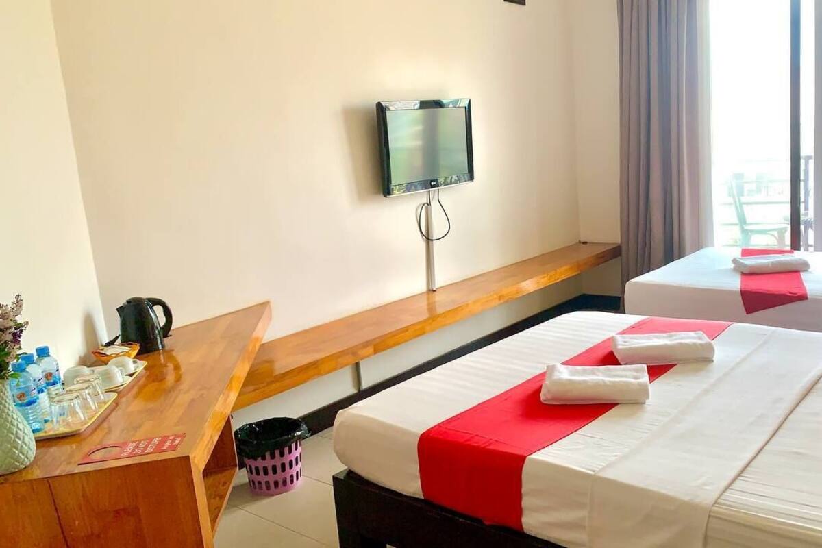 A hotel room with two beds, a TV mounted on the wall, a wooden bench, and a window with a view of the outside.