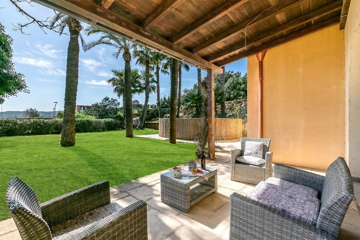 An outdoor patio with wicker furniture, a glass coffee table, and a view of a green lawn and palm trees.