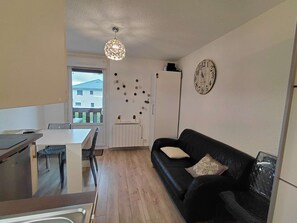 A compact living space with a dining area, a black leather sofa, a white refrigerator, and a wall clock. - Studio rénové grand confort 3 étoiles - Résidence Les Crêtets (Les Rousses)