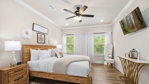 A bedroom with a wooden bed, bedside tables, a ceiling fan, a large window with curtains, and a mounted TV. - Discoverer - Ozark Mountain Retreat - Pets Allowed (Bella Vista)