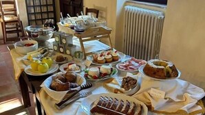 A dining table set with various food items, including fruits, pastries, and a cake, in a room with a window and a radiator. - Le Case di Cardellino - AgriSpa (Sclafani Bagni)