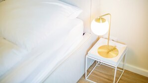 Una cama bien hecha con sábanas blancas, una mesita de noche pequeña con una lámpara blanca, y un piso de madera.