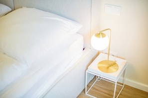 Una cama bien hecha con sábanas blancas, una mesita de noche pequeña con una lámpara blanca, y un piso de madera.