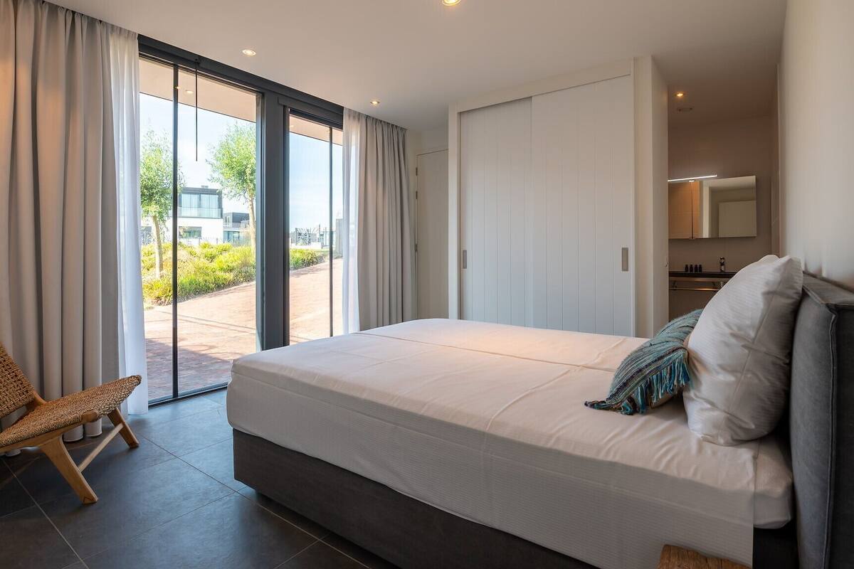 Une chambre à coucher moderne, dotée d’un grand lit, d’un fauteuil et offrant une vue sur l’extérieur grâce à une porte-fenêtre coulissante.