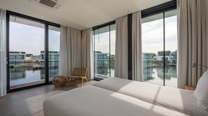 A modern hotel room with a large bed, a wicker chair, and a view of water and buildings. - Dormio Harbour Village (Arnemuiden)