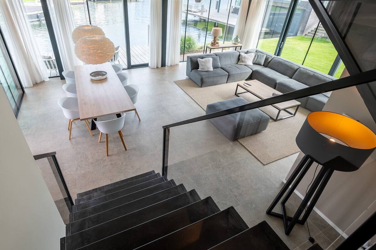 Un intérieur moderne avec un escalier menant à un espace de vie, un coin repas avec une table et des chaises, et une vue sur un plan d’eau grâce à de grandes fenêtres.
