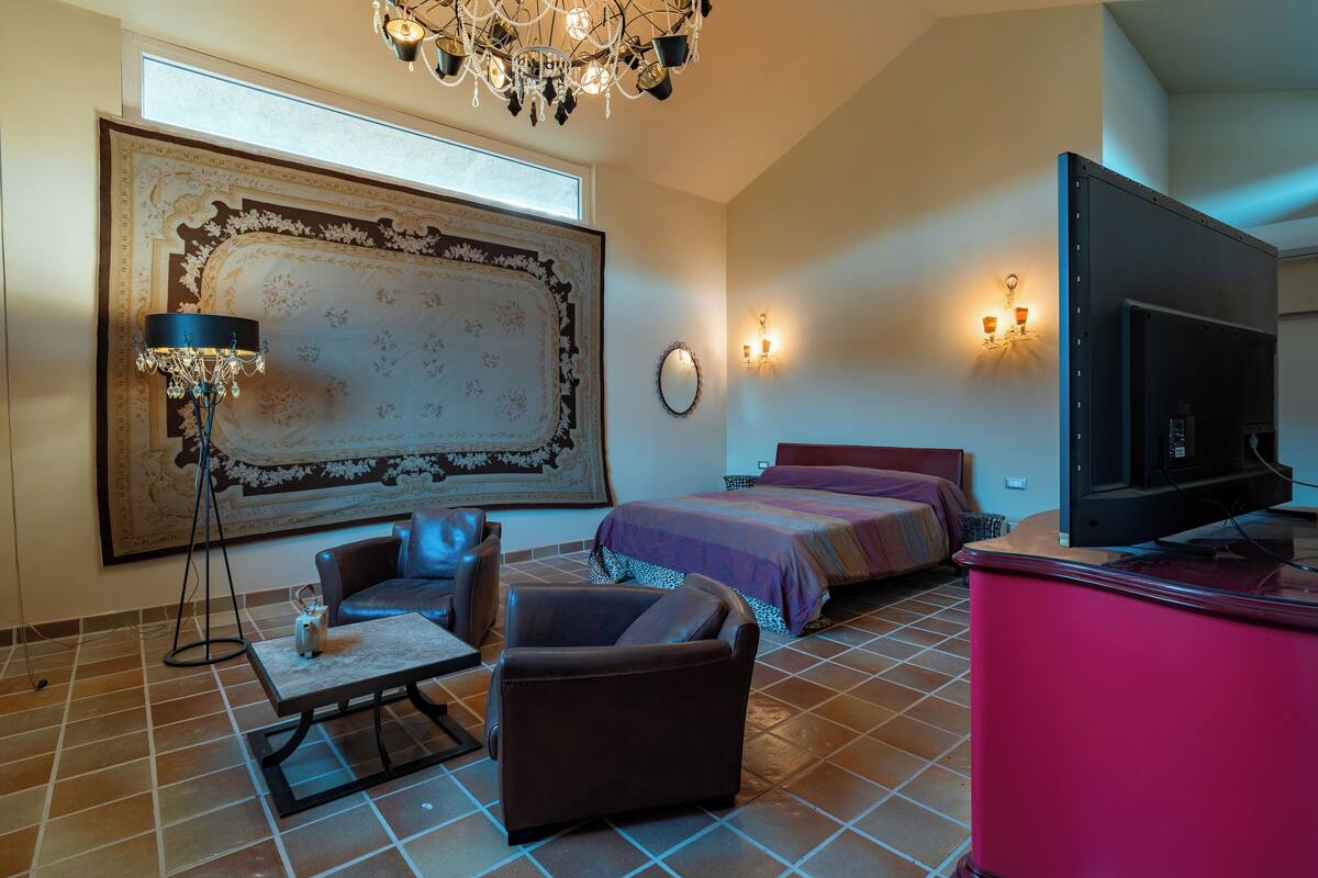 A hotel room with a large ornate rug, a modern chandelier, a flat-screen TV, and a bed with a purple bedspread.