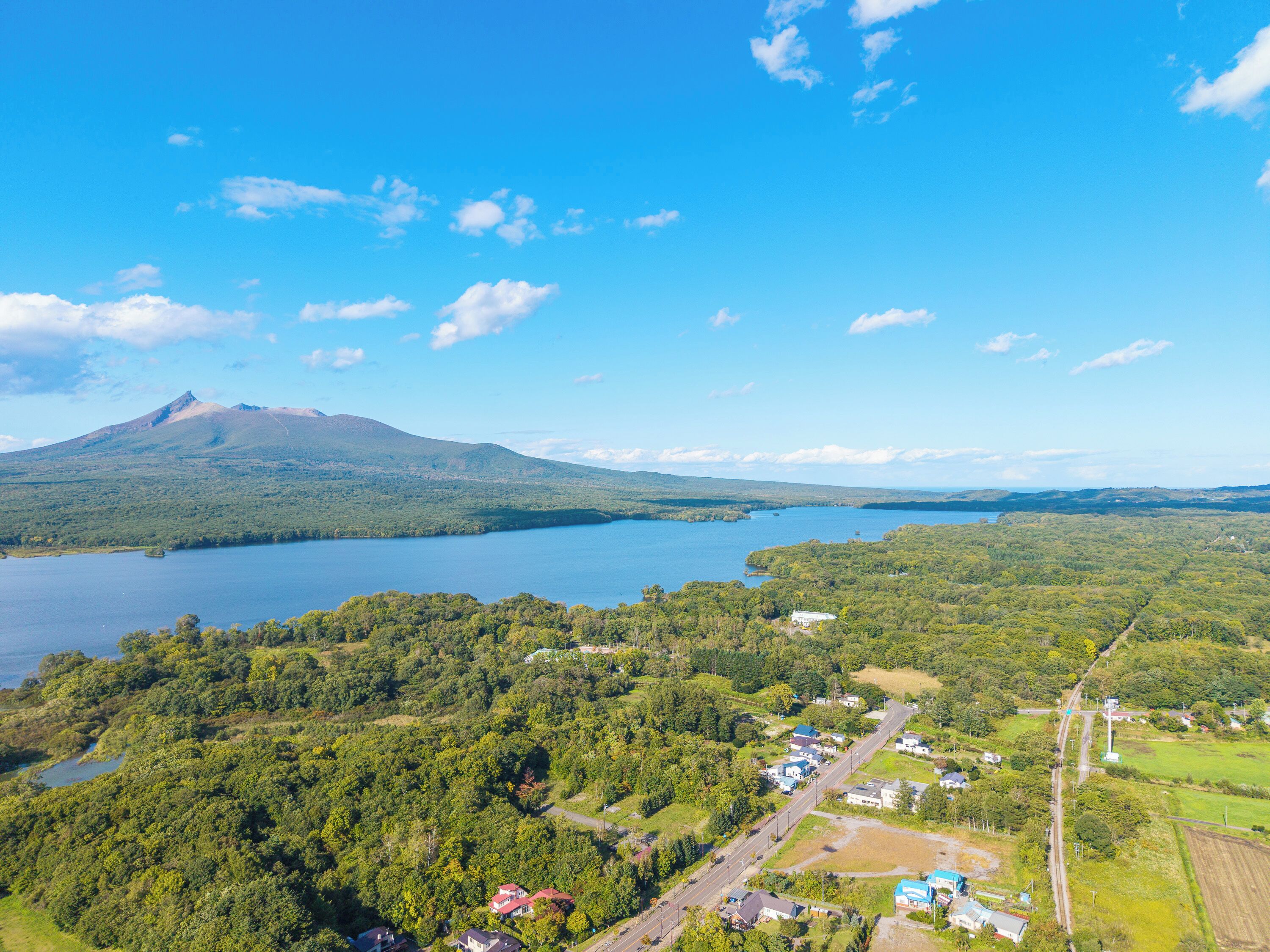 Une vue aérienne d’une zone au bord d’un lac, avec une montagne en arrière-plan, entourée de forêts denses et d’un petit village composé de maisons et de routes.