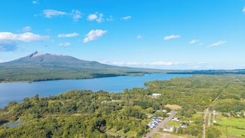 Une vue aérienne d’une zone au bord d’un lac, avec une montagne en arrière-plan, entourée de forêts denses et d’un petit village composé de maisons et de routes.