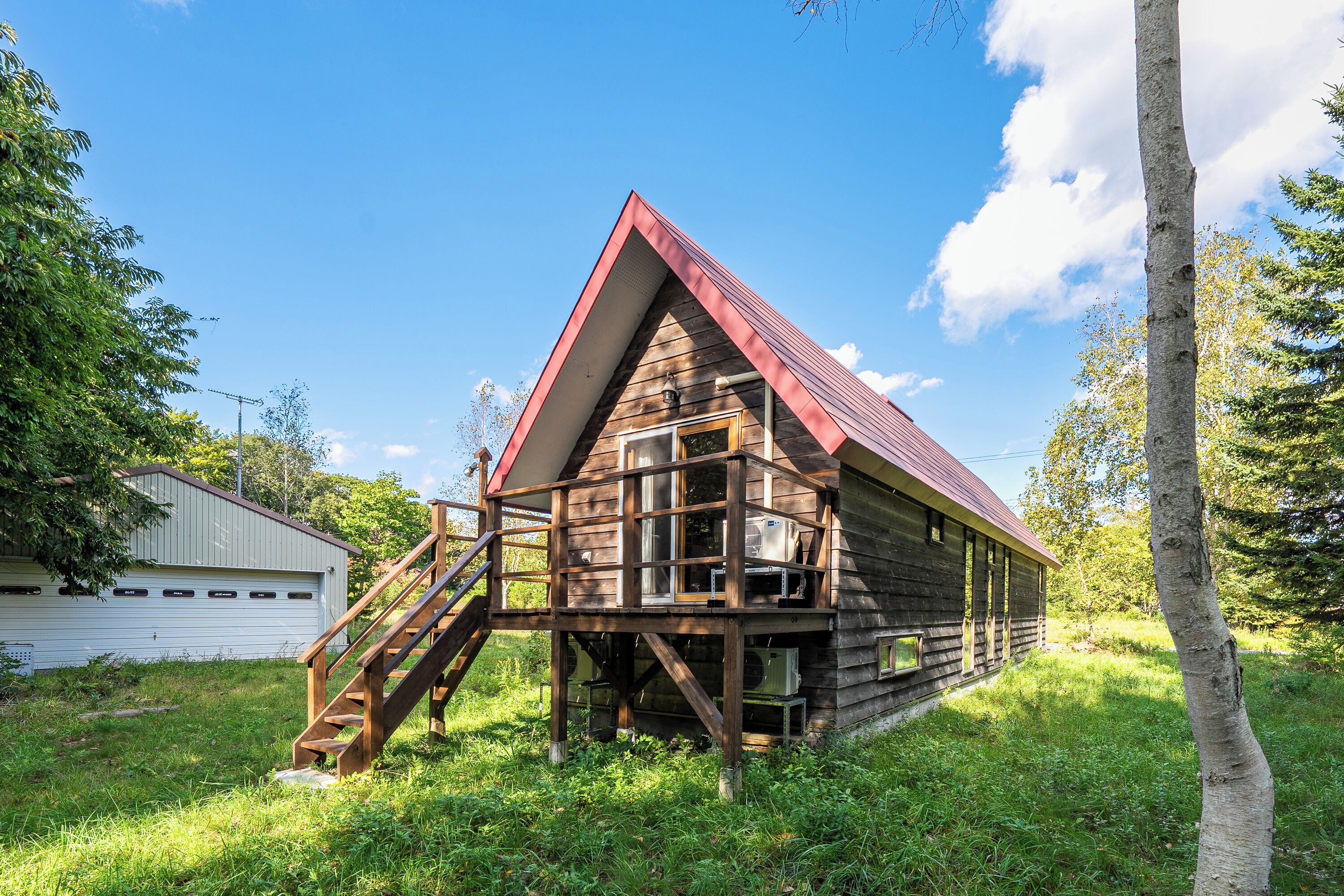 A rustic wooden cabin with a red roof, a wooden staircase leading to a deck, and a garage in the background.