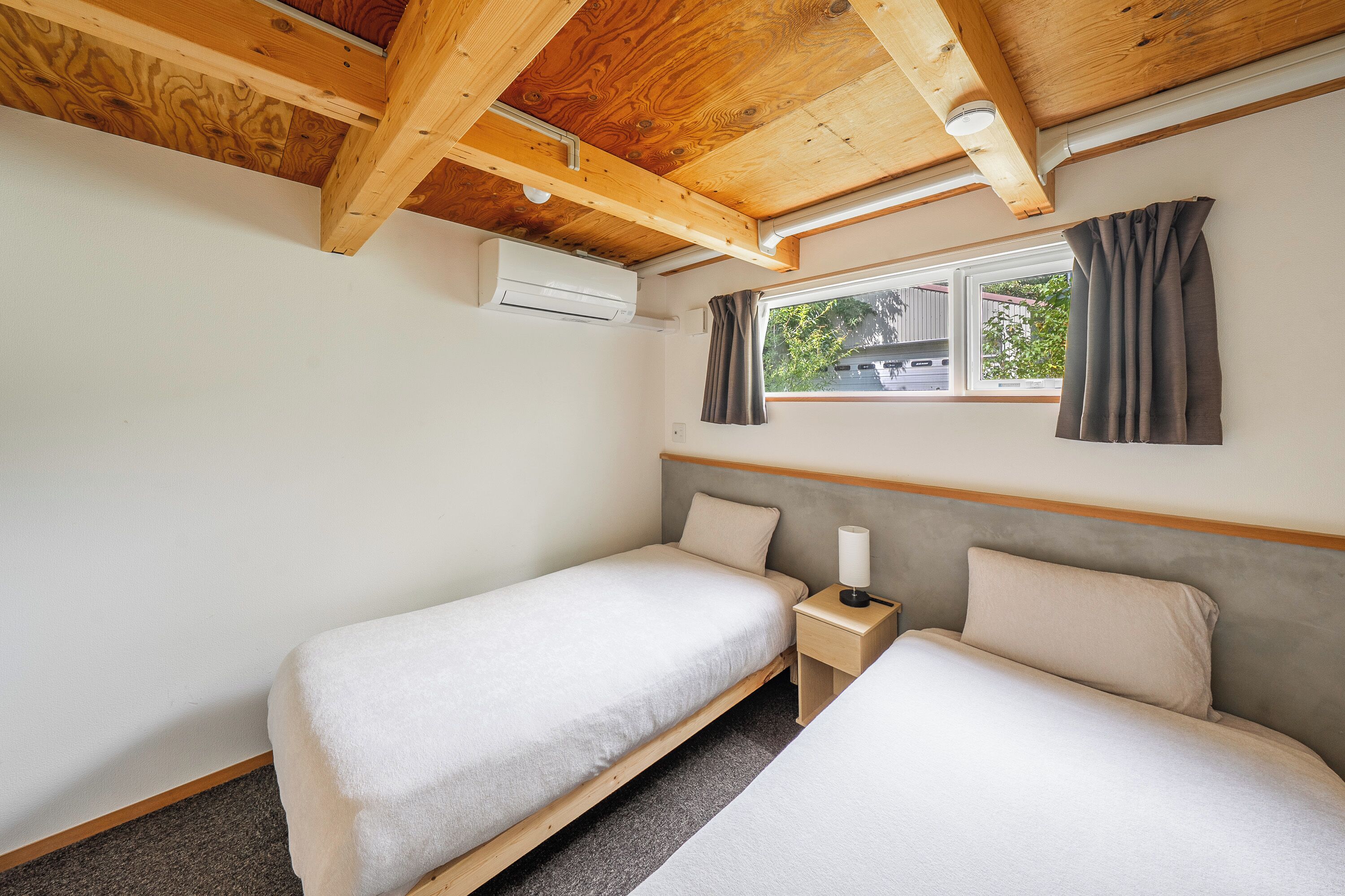 A small, clean room with two single beds, a window with curtains, and a wooden ceiling.