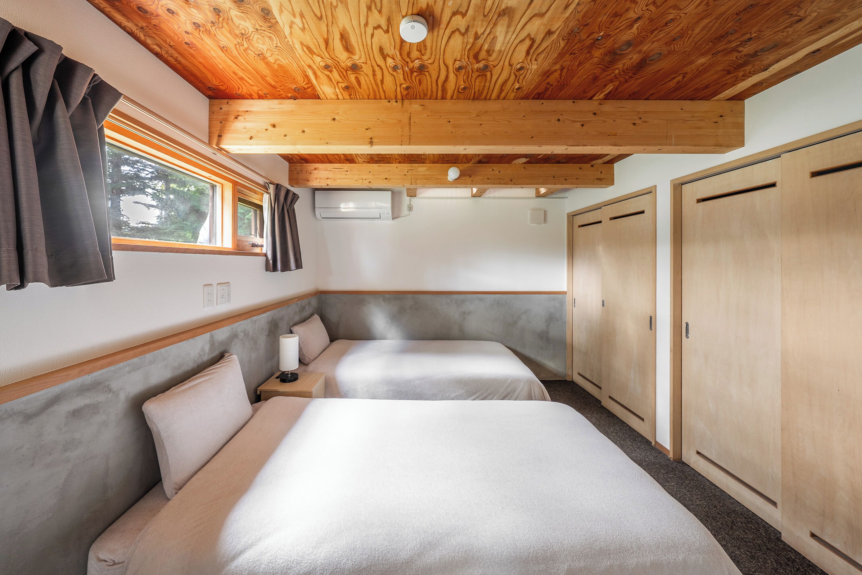 A cabin with two beds, wooden ceiling beams, a window with curtains, and a wardrobe.