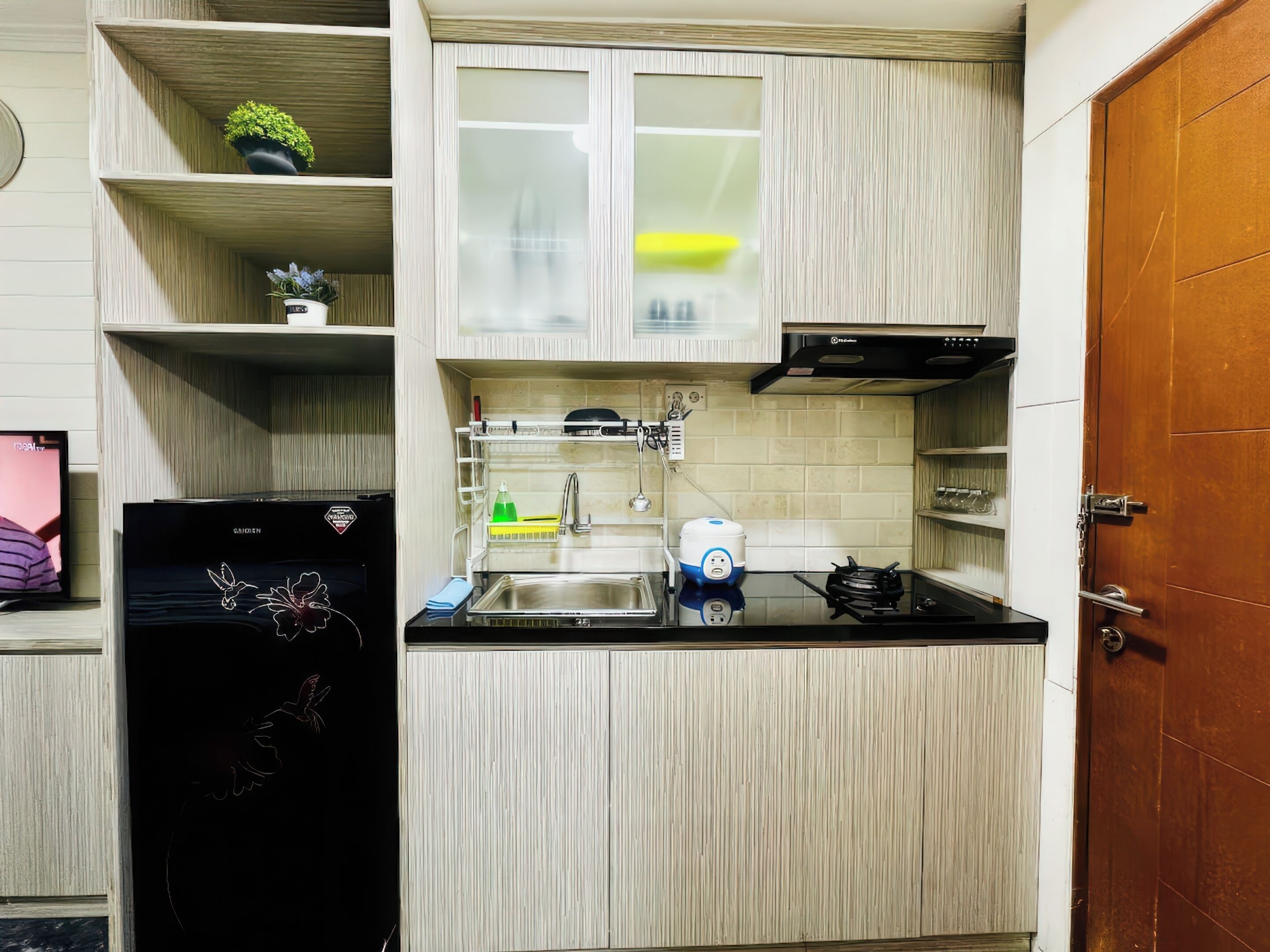 A compact kitchen with a refrigerator, sink, and a stove. There are shelves with plants and a window above the sink.