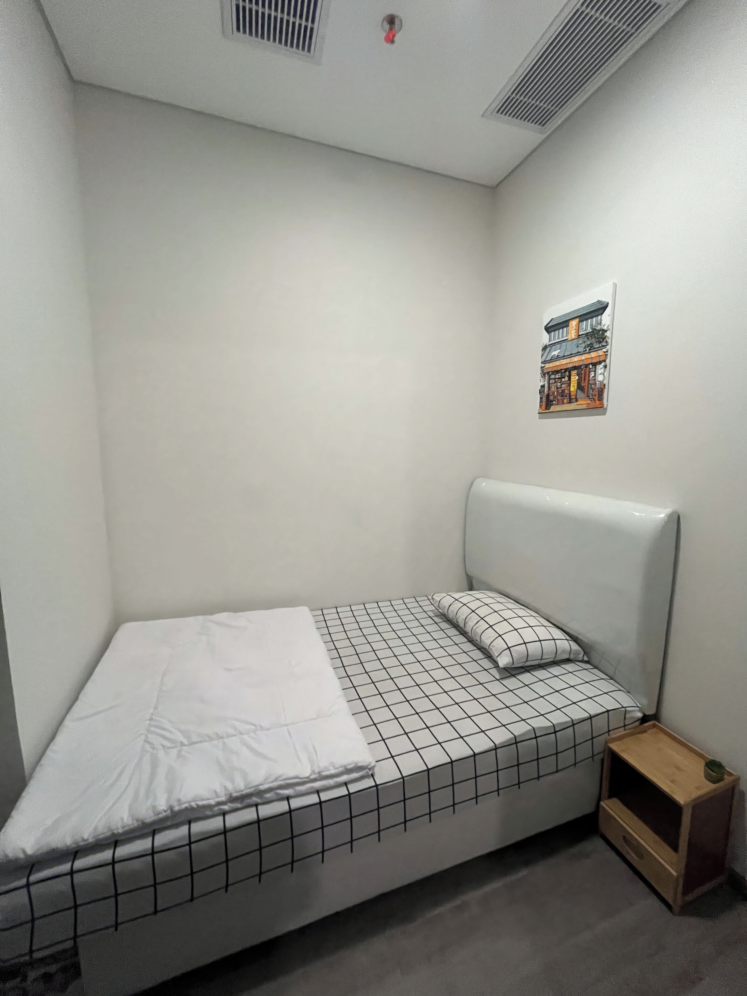 A small, neatly made bed with a white and black checkered bedspread and a single pillow. There is a small wooden nightstand with a green object on it. The room has white walls and a ceiling vent.