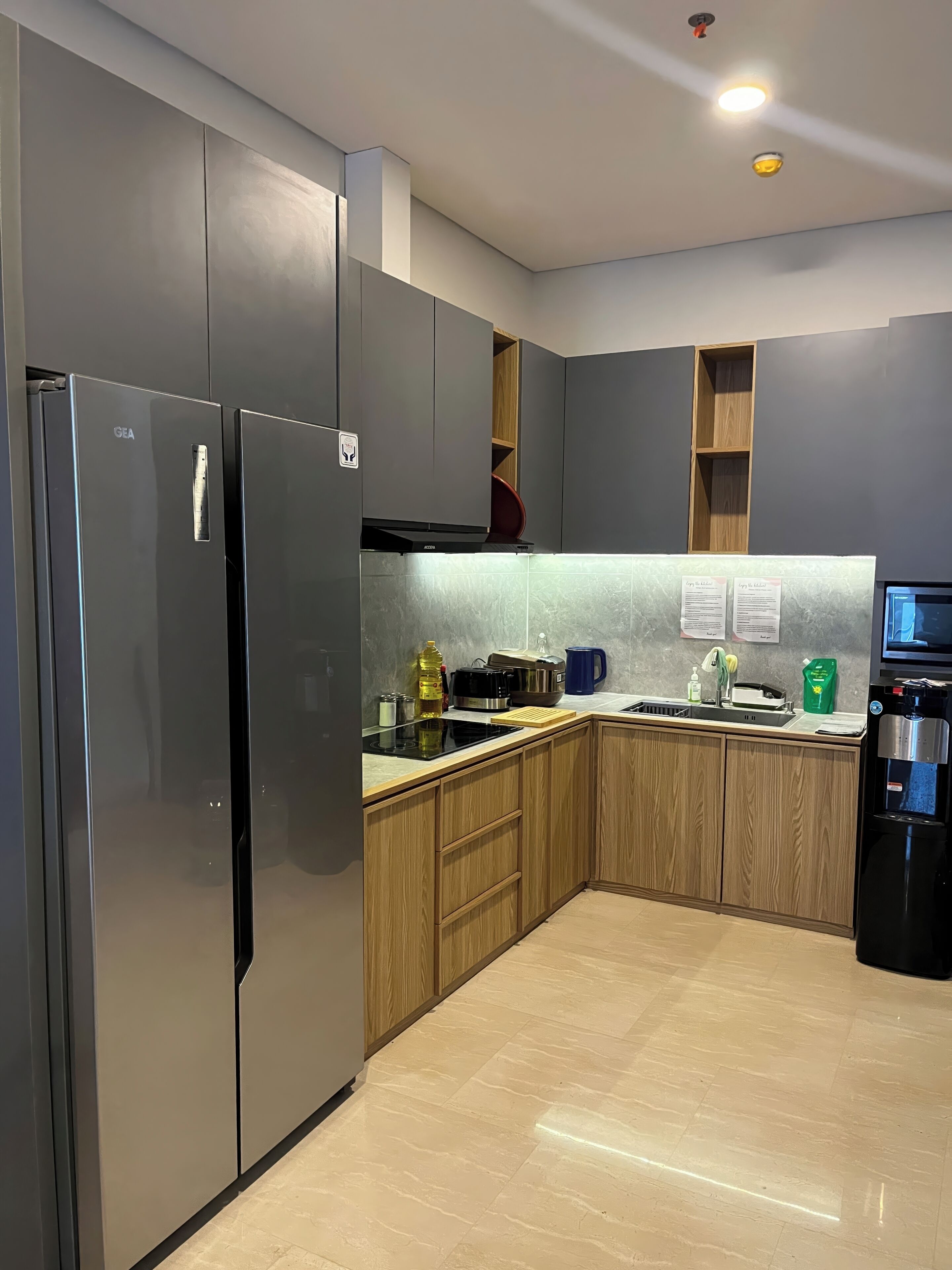 A modern kitchen with grey cabinets, a stainless steel refrigerator, wooden cabinets, a built-in oven, a sink, and a water dispenser.