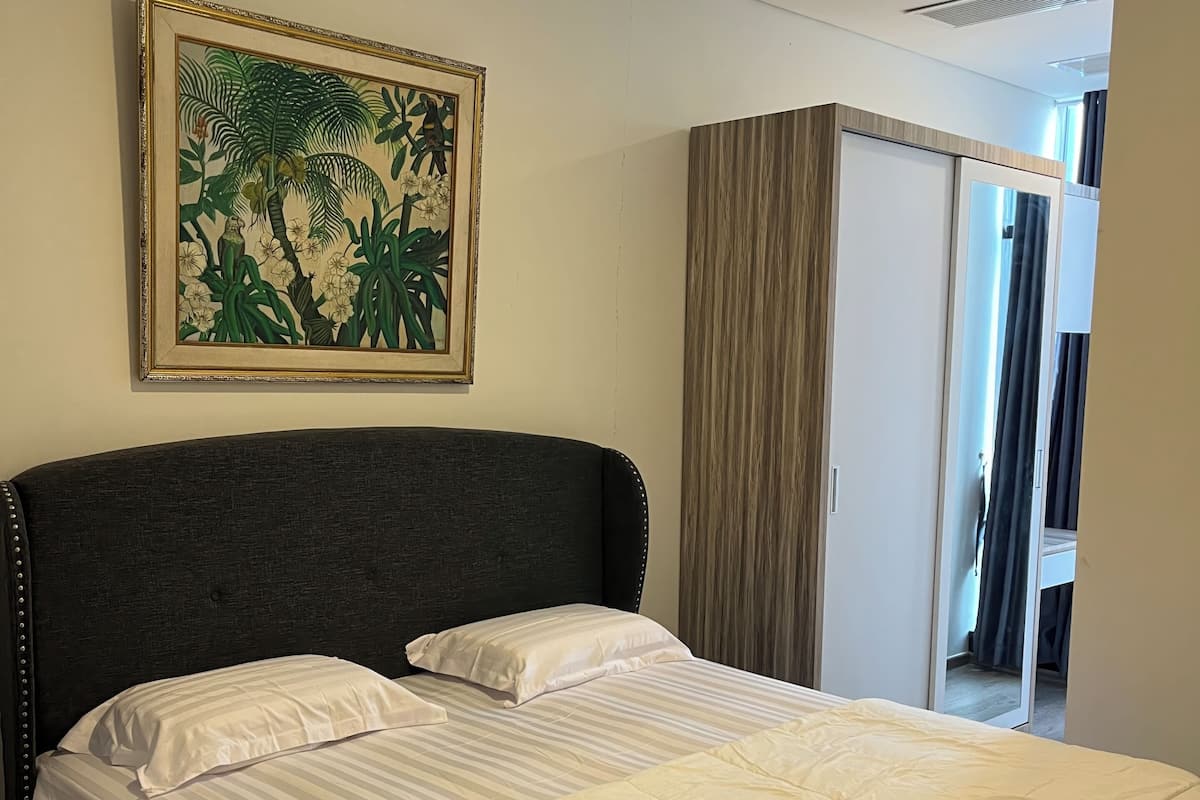 Cama bem arrumada com roupa de cama branca, cabeceira escura e quadro com arte de planta tropical na parede.