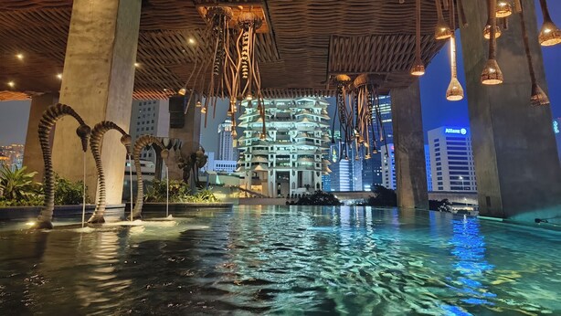 An indoor swimming pool with a wooden ceiling and hanging lights, surrounded by large concrete pillars and a cityscape view in the background. - Sudirman Suites Apartment Jakarta (Jakarta)