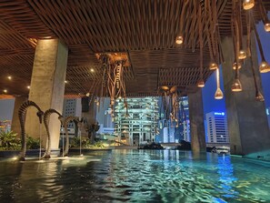 An indoor swimming pool with a wooden ceiling and hanging lights, surrounded by large concrete pillars and a cityscape view in the background. - Sudirman Suites Apartment Jakarta (Jakarta)