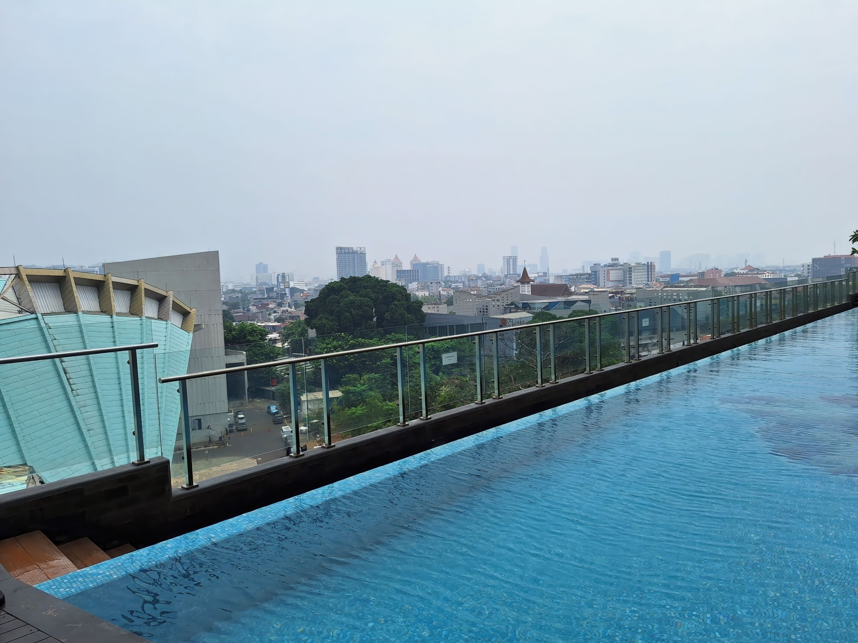 A rooftop swimming pool with a glass balustrade, overlooking a cityscape with various buildings and a large tree.