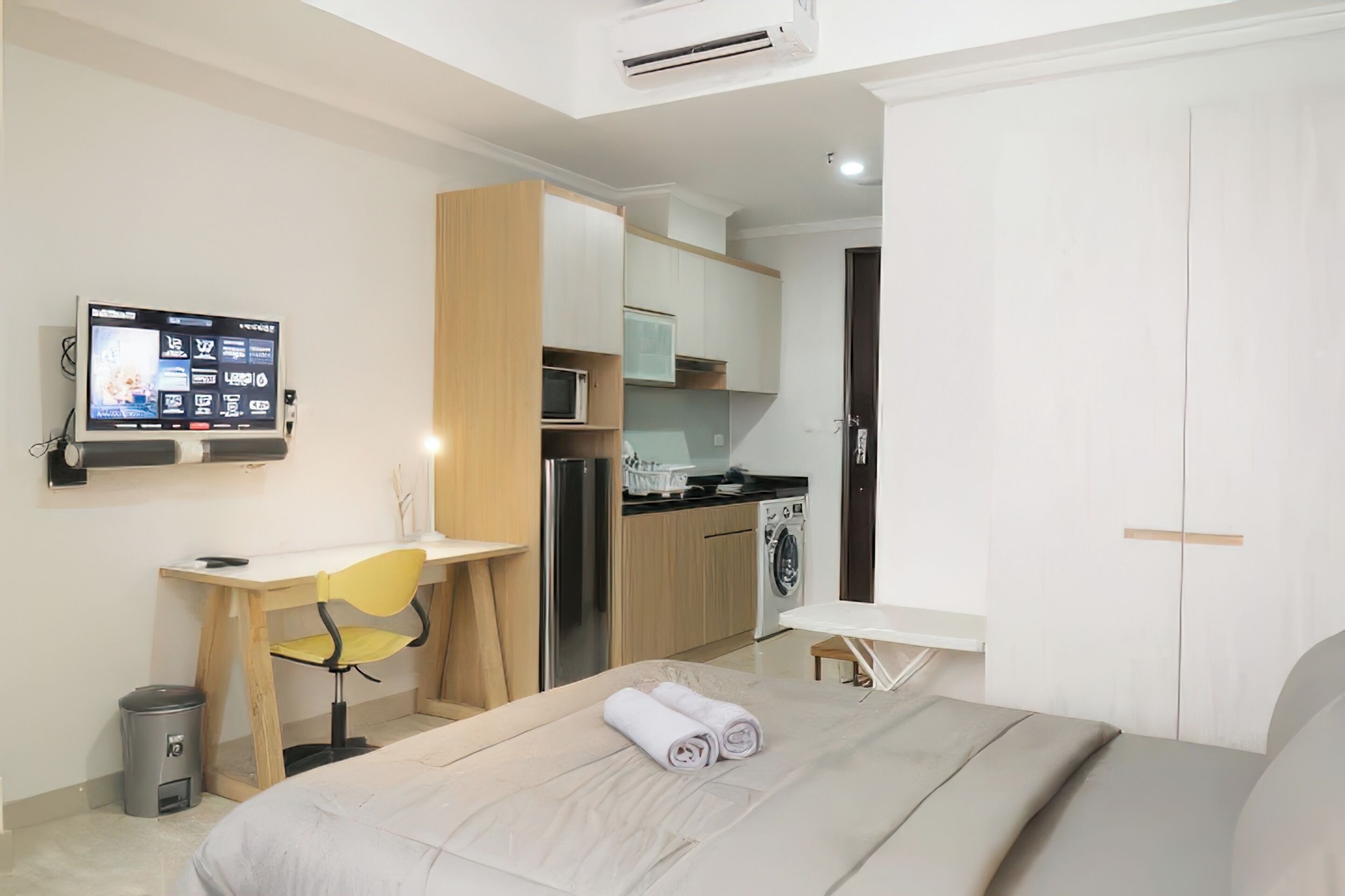 A modern hotel room with a bed, a desk, a chair, a TV, a microwave, a washing machine, and a small table.
