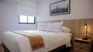 Un letto rifatto con lenzuola bianche e una coperta beige, un comodino con una lampada e un quadro con vista sulla città appeso al muro.