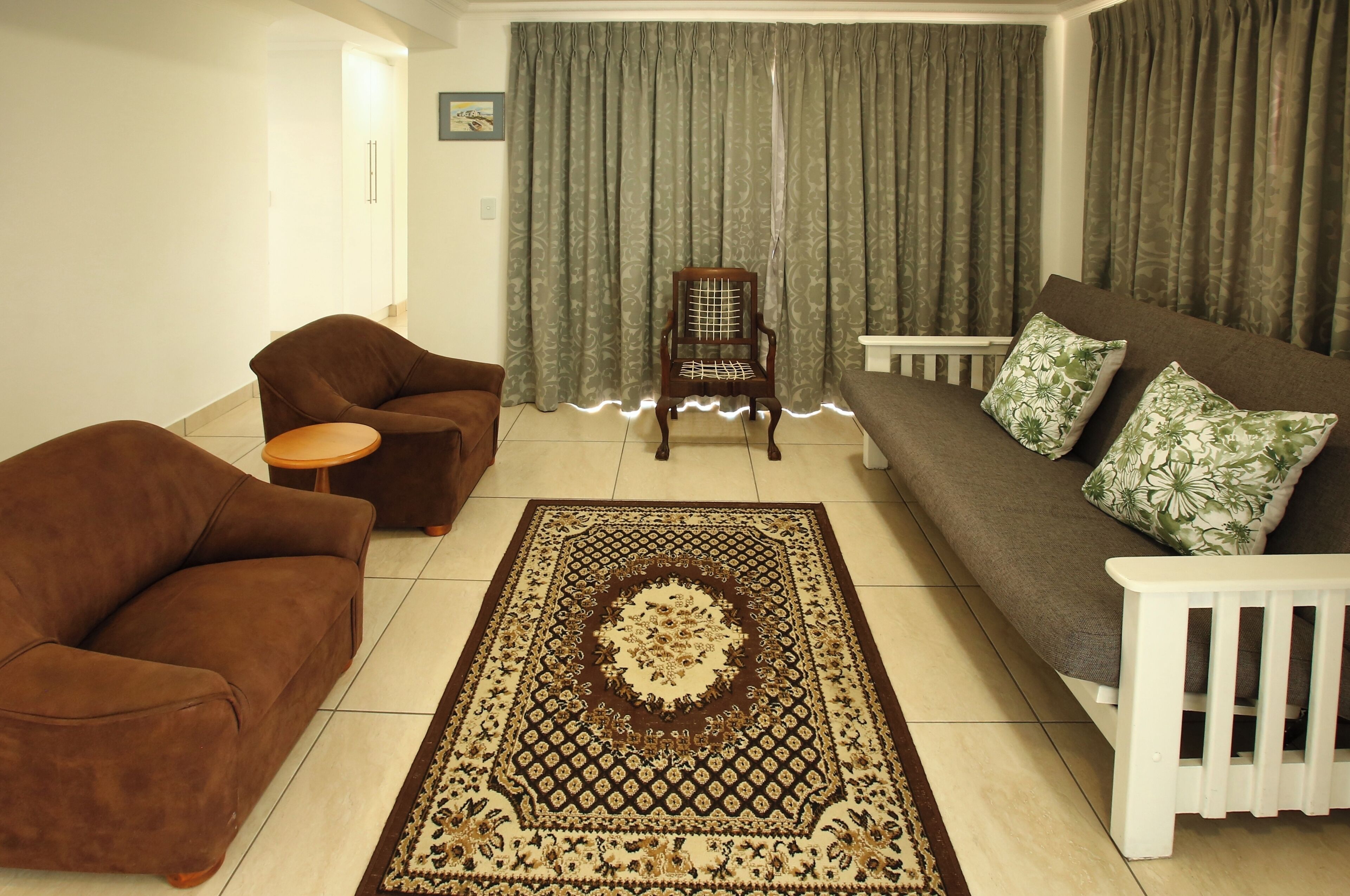 A living room with a brown sofa, a brown armchair, a wooden chair, a patterned rug, and green curtains.