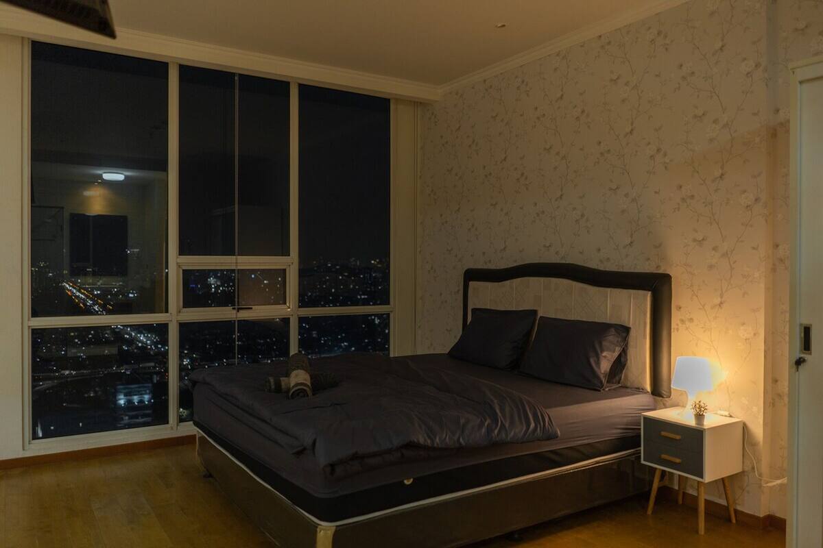 Un dormitorio con un ventanal grande, una cama, una mesita de noche con lámpara y vista a un paisaje urbano nocturno.