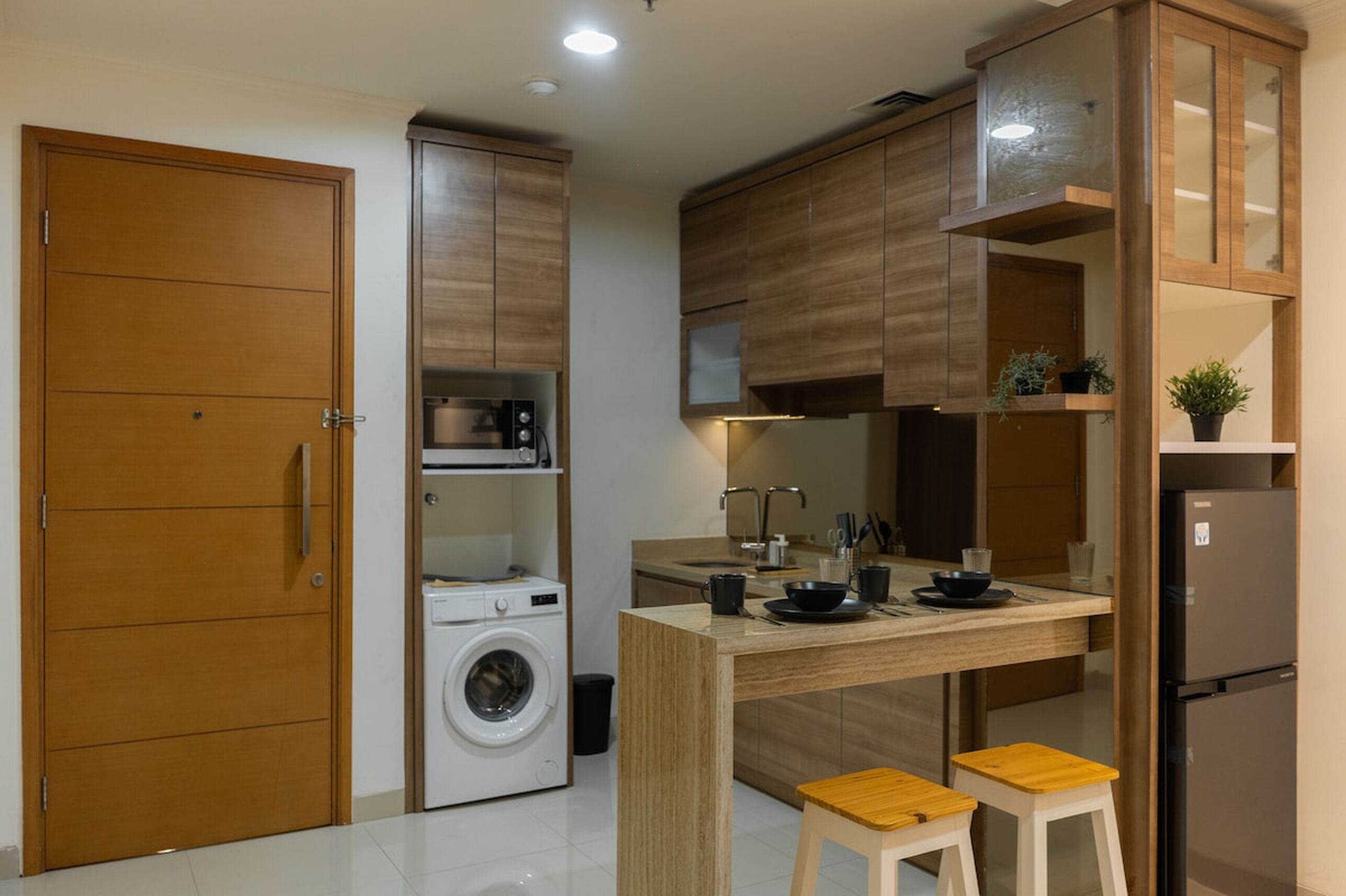 A compact kitchen with wooden cabinets, a washing machine, a dining area with stools, and a refrigerator.