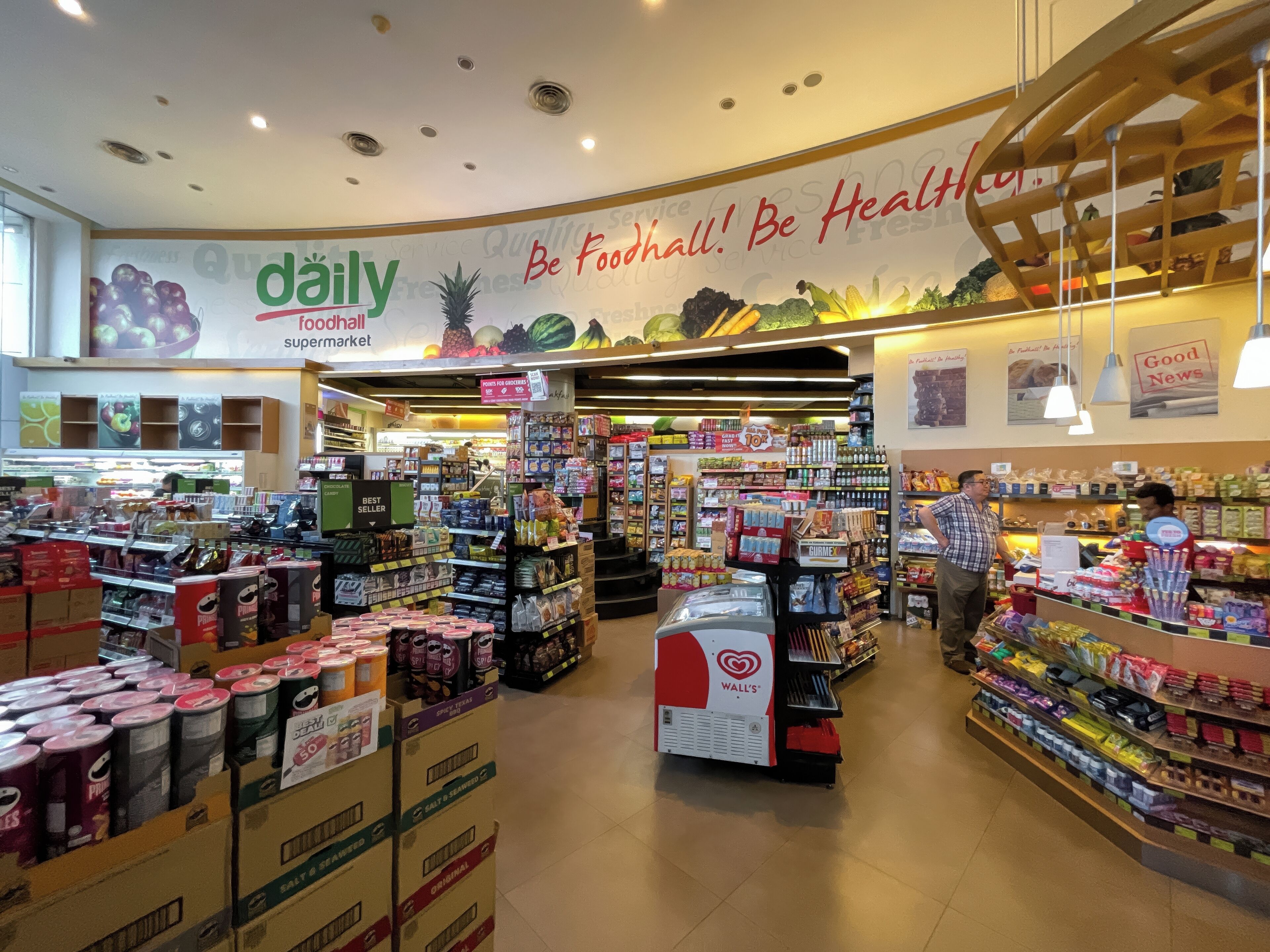 Interior de supermercado com diversos corredores repletos de produtos, um sinal chamativo com a palavra 'diário' e o slogan 'Be Foodhall! Be Healthy!' na parede.