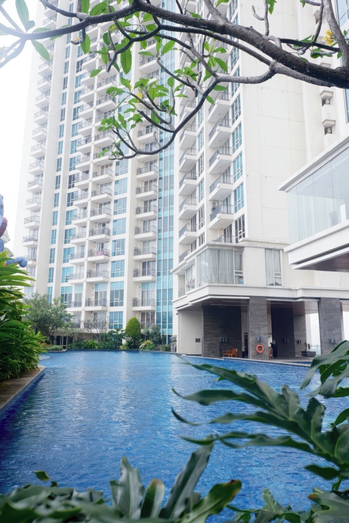 A modern building with a swimming pool, surrounded by greenery and a tall residential building in the background.