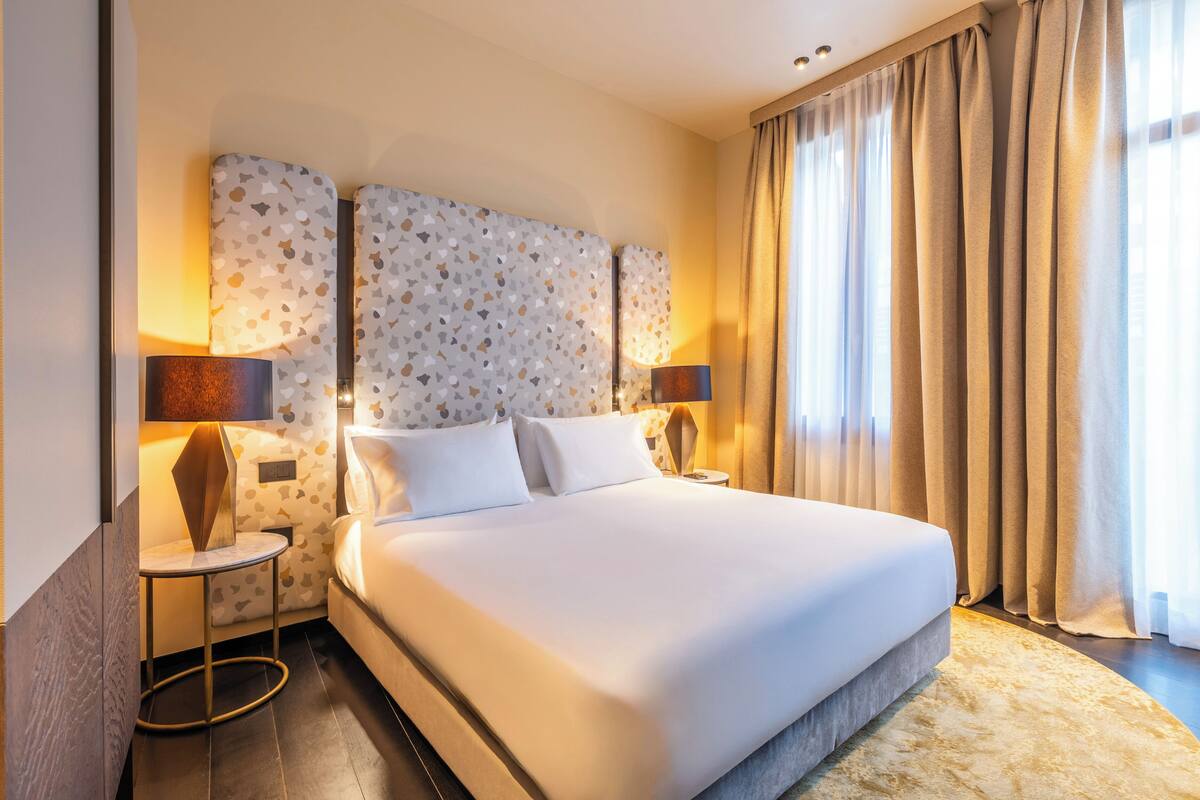 A hotel room with a large bed, two bedside tables with lamps, a headboard with a leaf pattern, and a view of the outdoors through curtains.