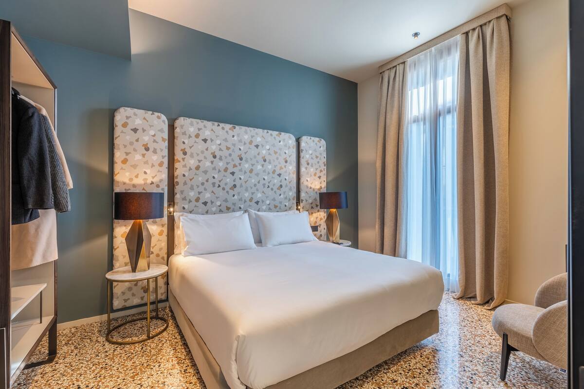 A hotel room with a large bed, two bedside lamps, a small side table, a chair, and a wardrobe.