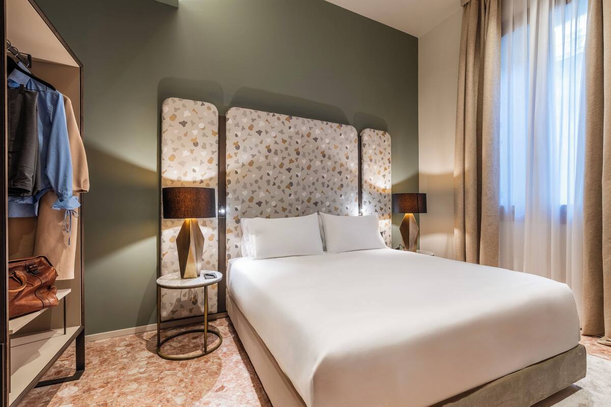 A hotel room with a large bed, a wardrobe, a small table with a lamp, and a view of the outdoors through the window.