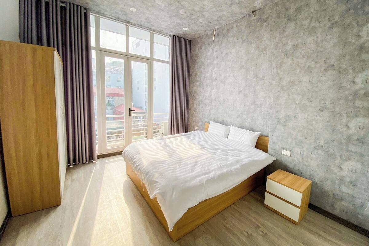 A modern bedroom with a large bed, wooden nightstand, and a view through a large window.