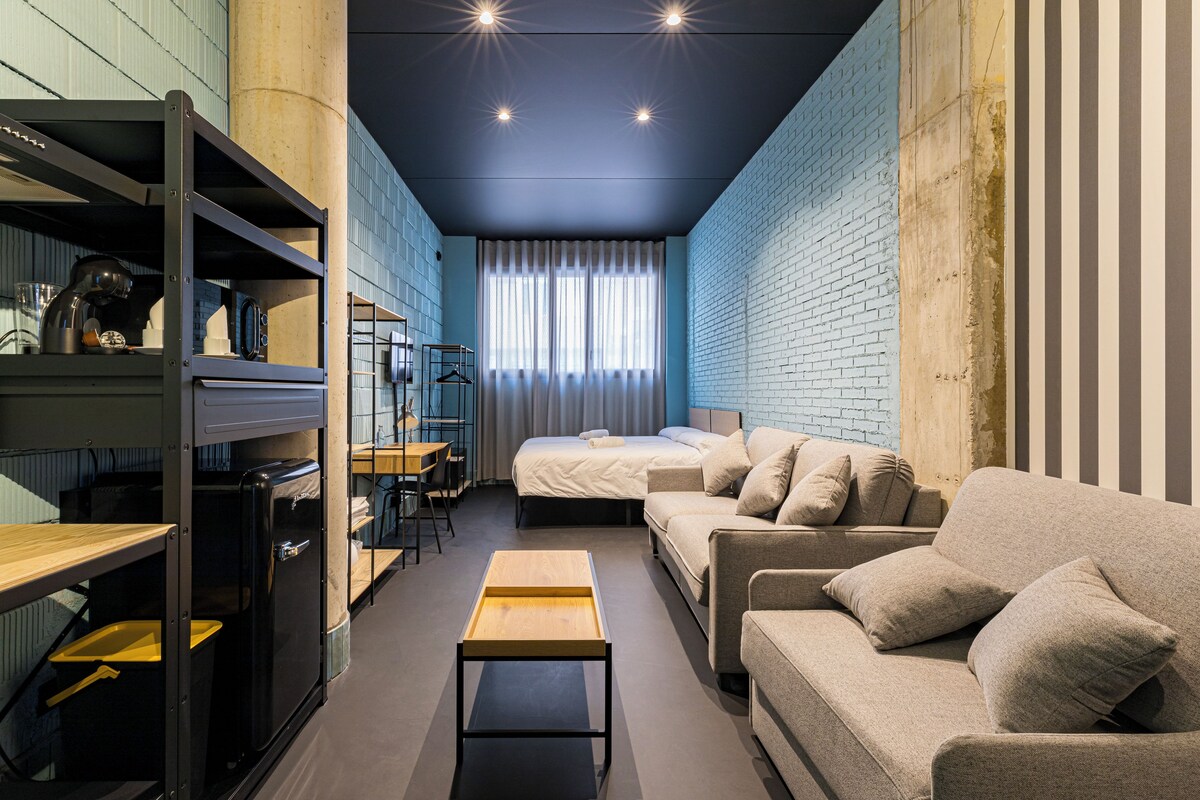 A compact hotel room with a bed, two sofas, a small table, and built-in shelves.