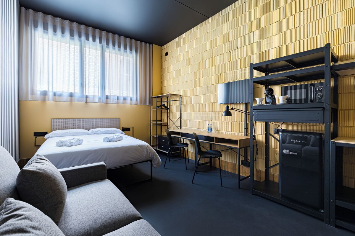 A modern hotel room with a bed, a desk, a chair, and a minibar.