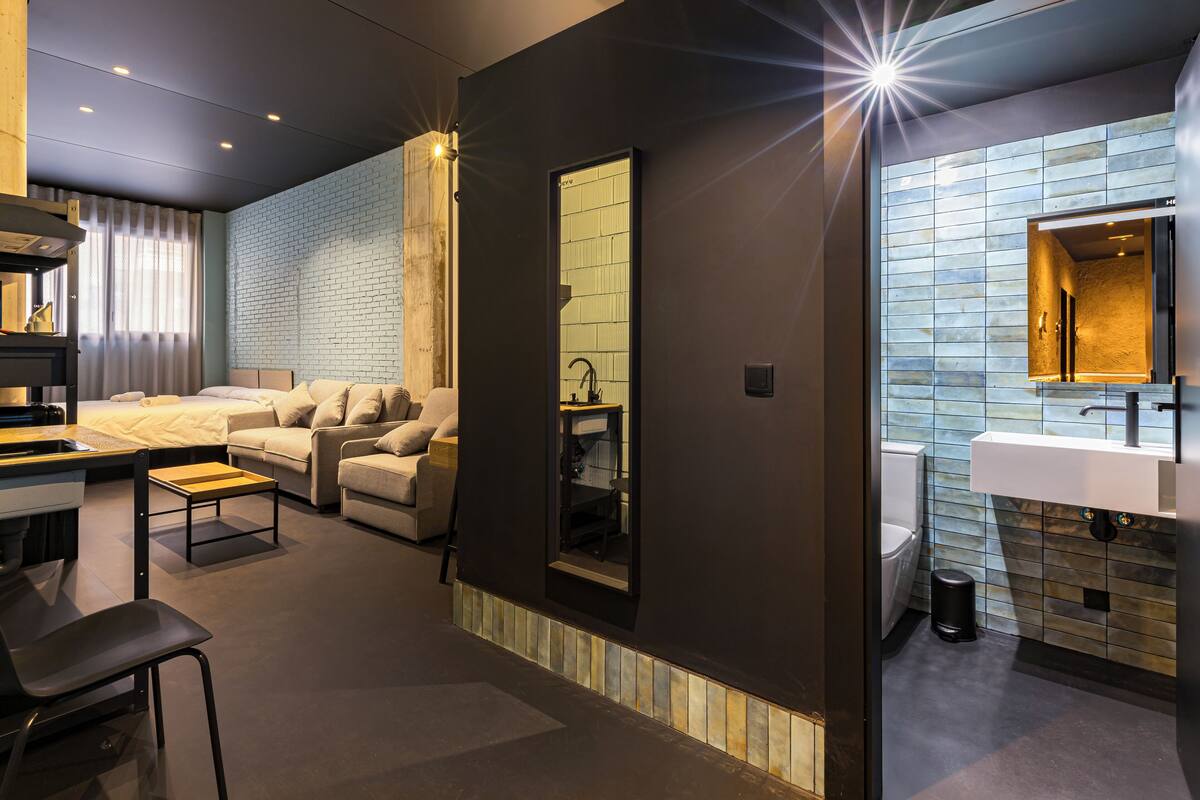 A modern hotel room with a sofa, a small table, a desk, a mirror, and a bathroom with a sink and toilet.