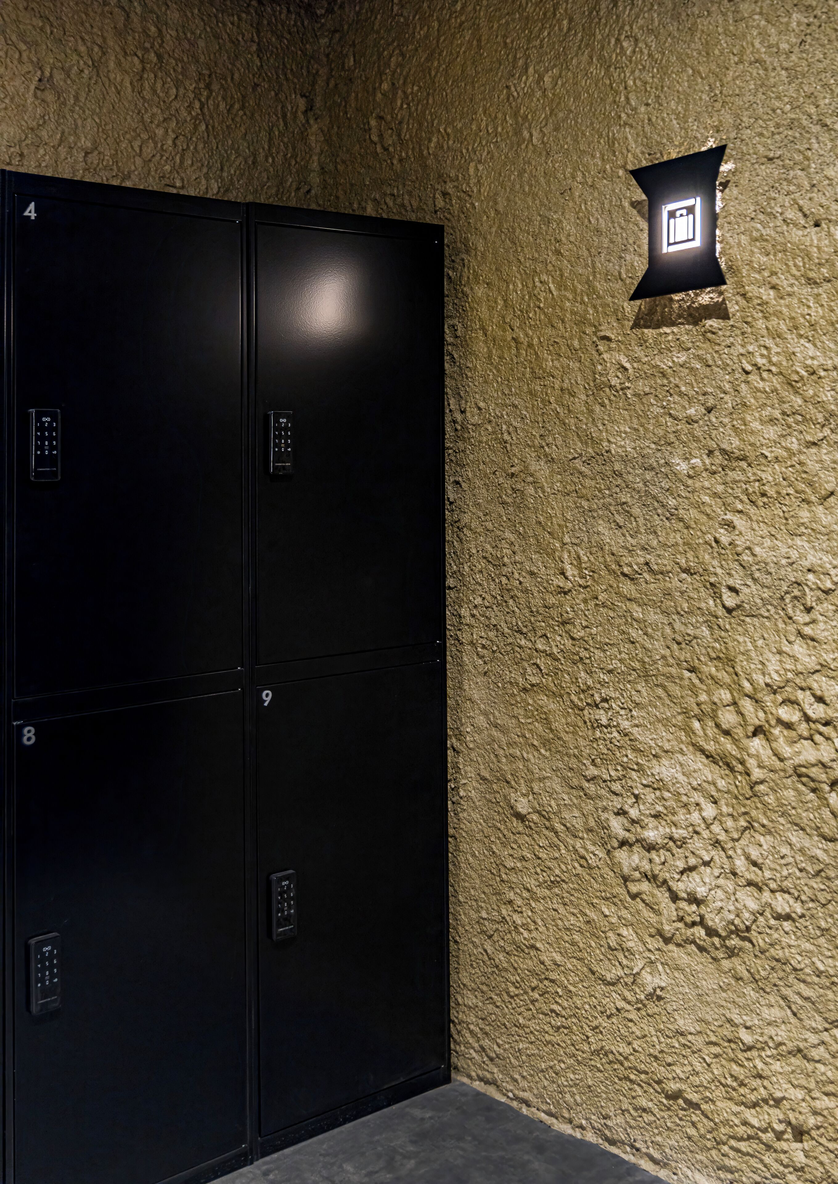 A set of four black lockers with numbered doors, a textured beige wall, and a small illuminated sign above.