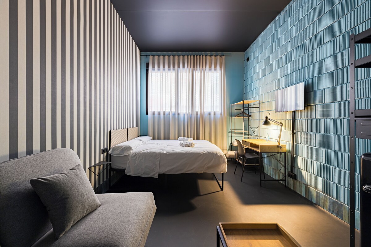 A modern hotel room with a bed, a sofa, a desk, and a TV mounted on the wall.