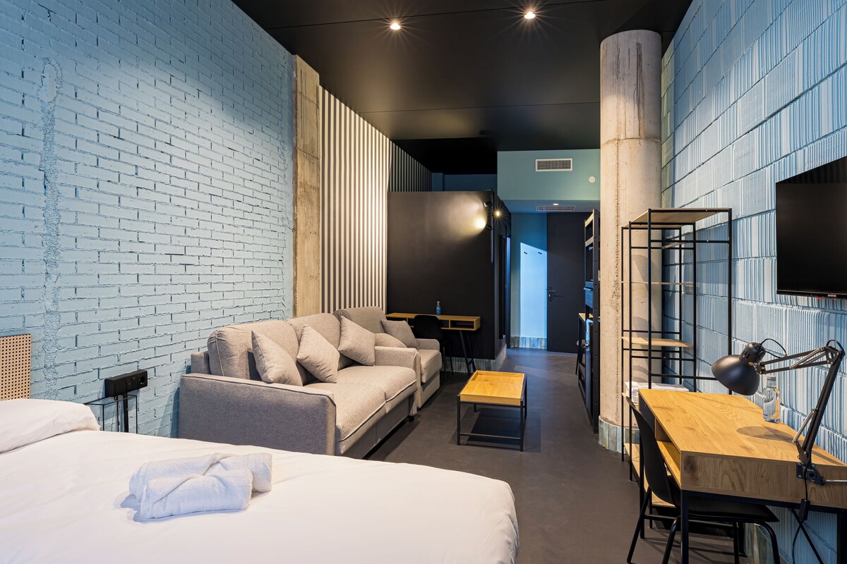A modern hotel room with a brick wall, a sofa, a bed, a desk, and a chair.