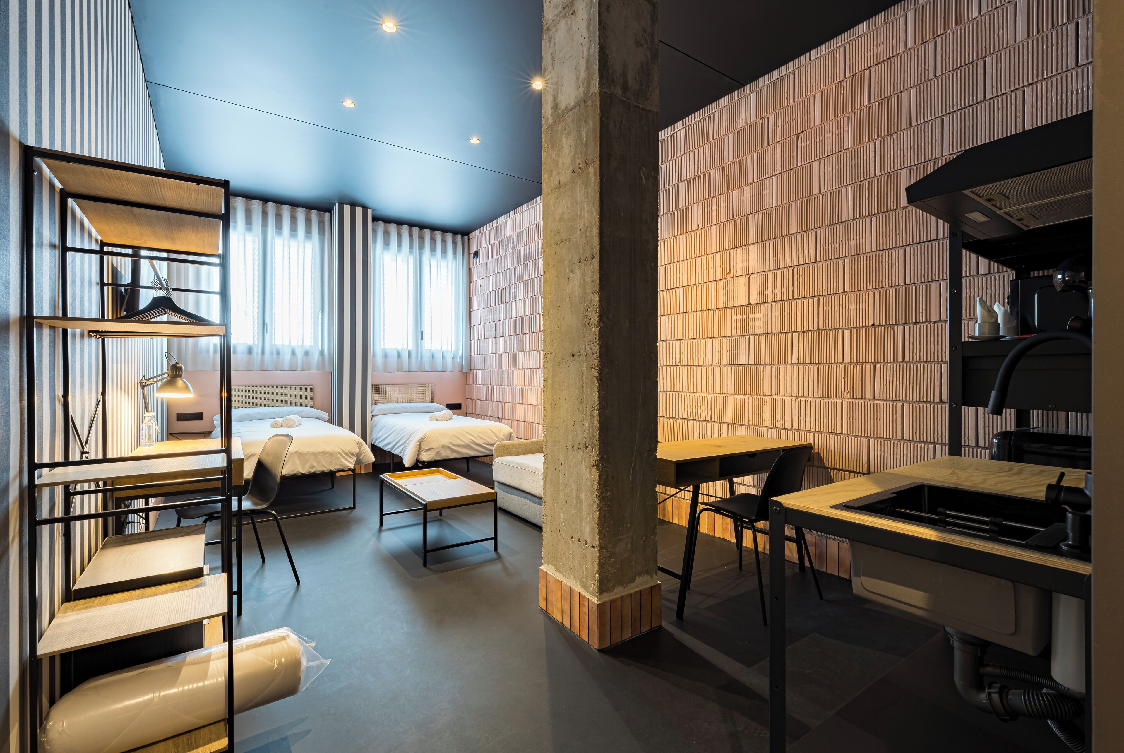 Habitación de hotel moderna con cama, escritorio, silla y una mesita. Se aprecia una ventana con cortinas, una pared con diseño de ladrillo y un techo con iluminación empotrada.
