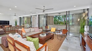 A spacious living room with a large sofa, coffee table, and chairs. There is a TV, a fireplace, and large windows offering a view of the outdoors.