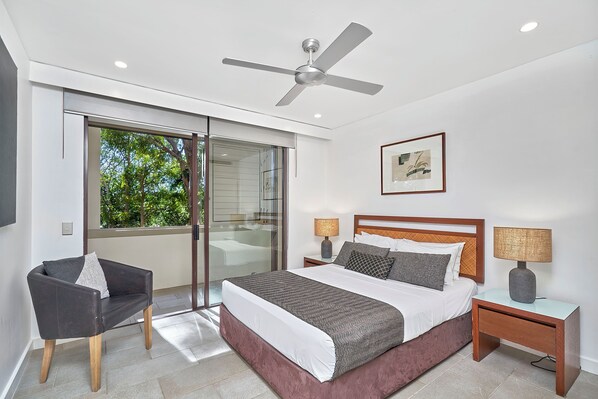 A modern bedroom with a large bed, a ceiling fan, a glass door leading to a balcony, and two bedside tables with lamps.