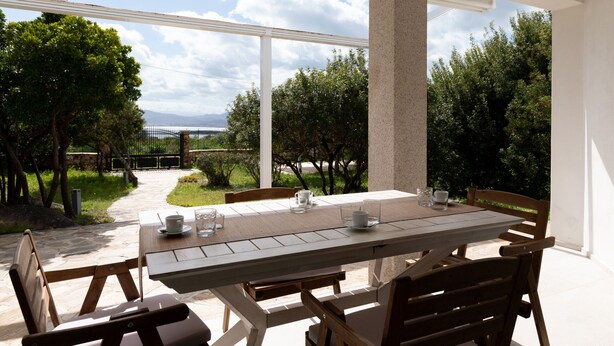 A patio with a table set for two, surrounded by wooden chairs, overlooking a garden with trees and a pathway. - Villetta Otis by Wonderful Italy (San Teodoro)