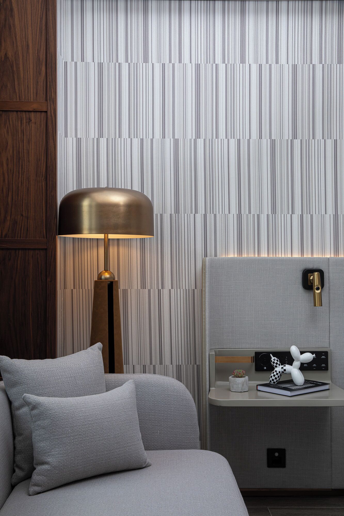 A modern hotel room with a striped wallpaper, a wooden panel wall, a bed with grey bedding, a bedside lamp, a nightstand with a book and a decorative item, and a control panel.