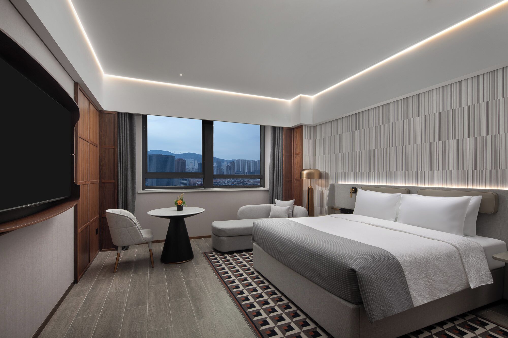 A modern hotel room with a large bed, a seating area, a TV, and a city view.
